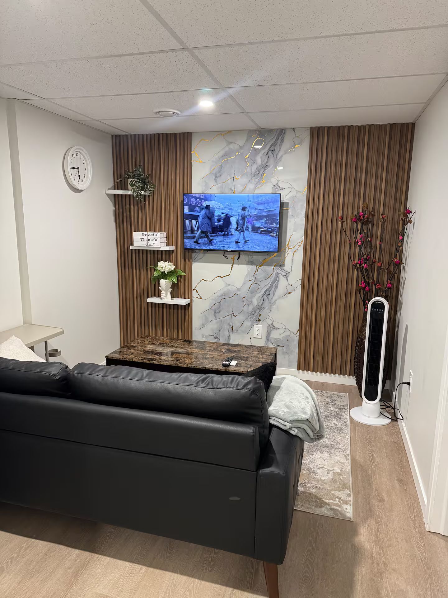 A modern living room with a black leather sofa, a flat-screen TV mounted on a marble wall, a small white clock, and a decorative plant.