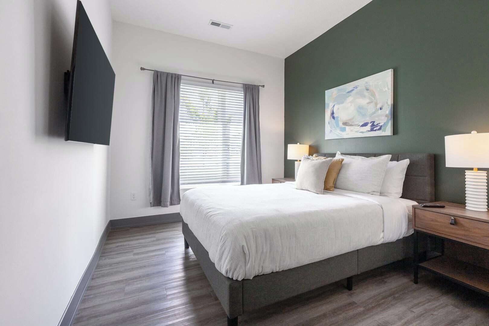 A hotel room with a bed, a bedside table with a lamp, a TV mounted on the wall, and a window with curtains.