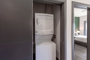 Un área de lavandería compacta con lavadora y secadora, una puerta que da a un dormitorio con cama y mesita de noche.