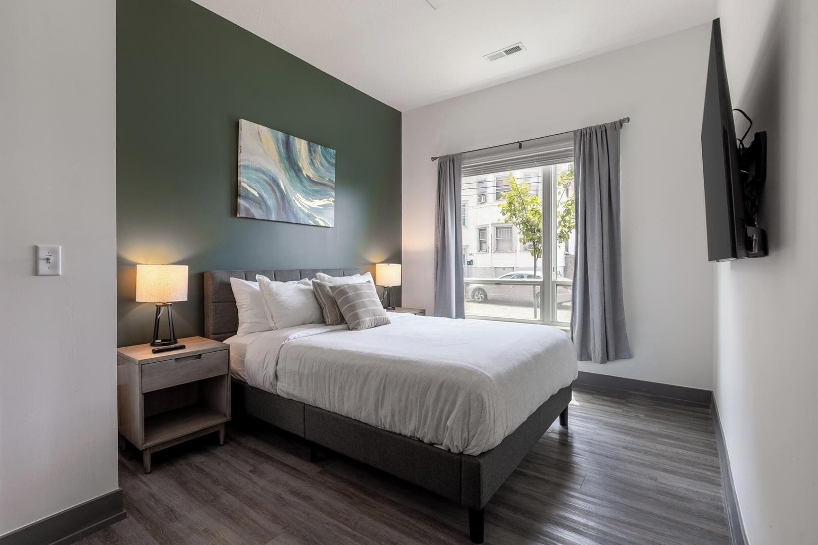 Une chambre d’hôtel avec un lit, des lampes de chevet, un tableau au mur, une fenêtre avec des rideaux et un téléviseur à écran plat fixé au mur.