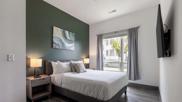A hotel room with a bed, bedside lamps, a painting on the wall, a window with curtains, and a flat-screen TV mounted on the wall. - Landing Amazing 2bd, Courtyard, Clubhouse (Dayton)