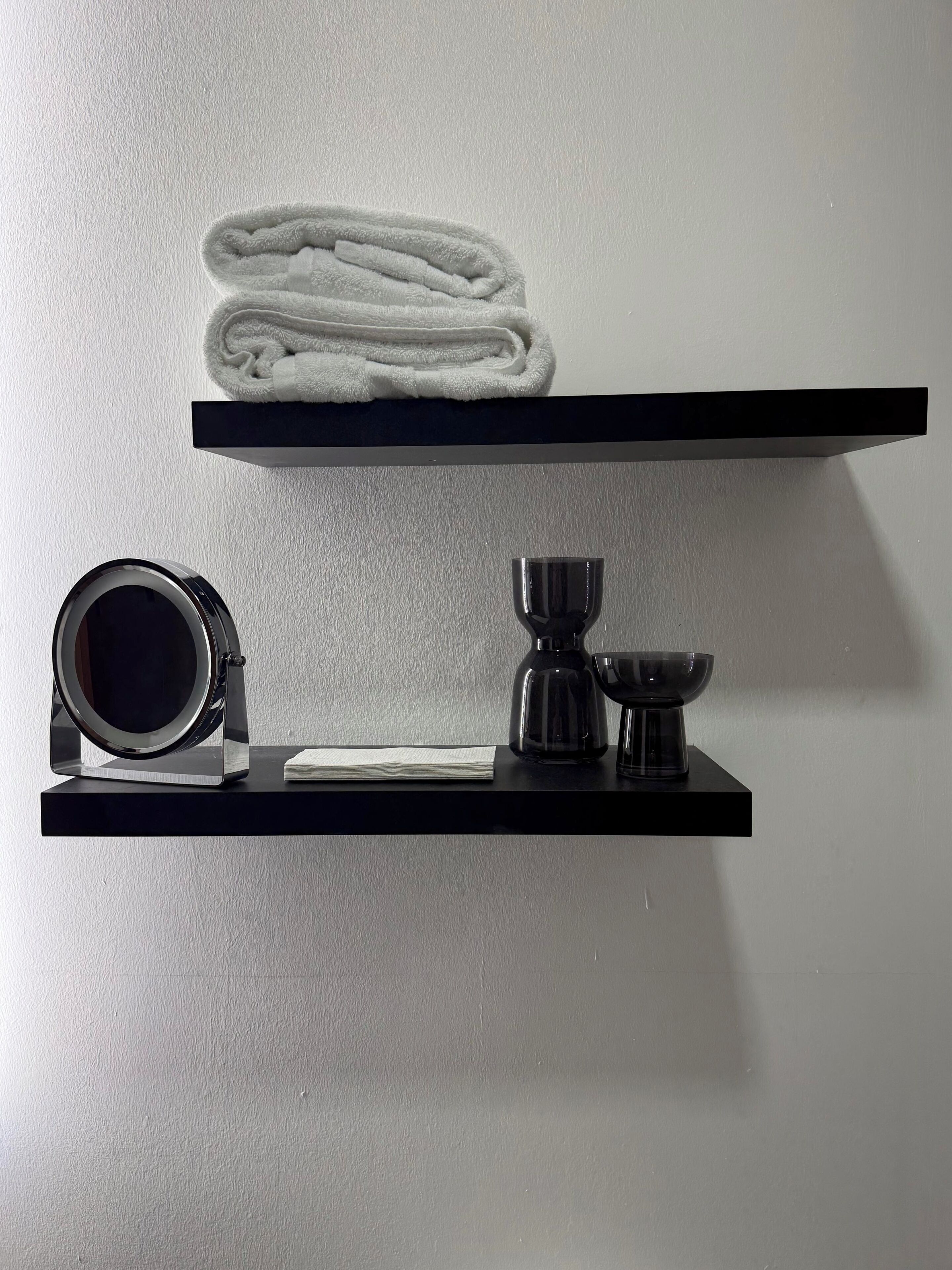 Deux étagères noires adossées à un mur blanc, l’une contenant une serviette blanche pliée et l’autre, un appareil photo, un verre et un petit bol.