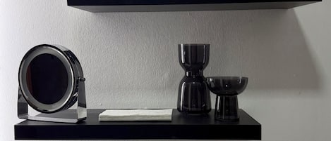 Two black shelves against a white wall, one holding a folded white towel and the other holding a camera, a glass, and a small bowl.