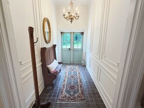 A hallway with a bench, a chandelier, a patterned rug, and a door with a view of greenery outside.