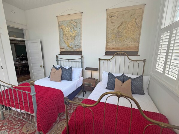 A room with two beds, a red knitted blanket, a map on the wall, and a window with shutters.