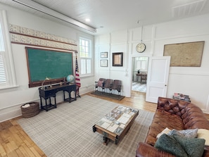 A room with a large green chalkboard, a wooden desk with a globe, a brown leather couch, and a patterned coffee table.