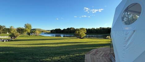 Una carpa con ventanal circular, una terraza de madera y vistas a un río y árboles.