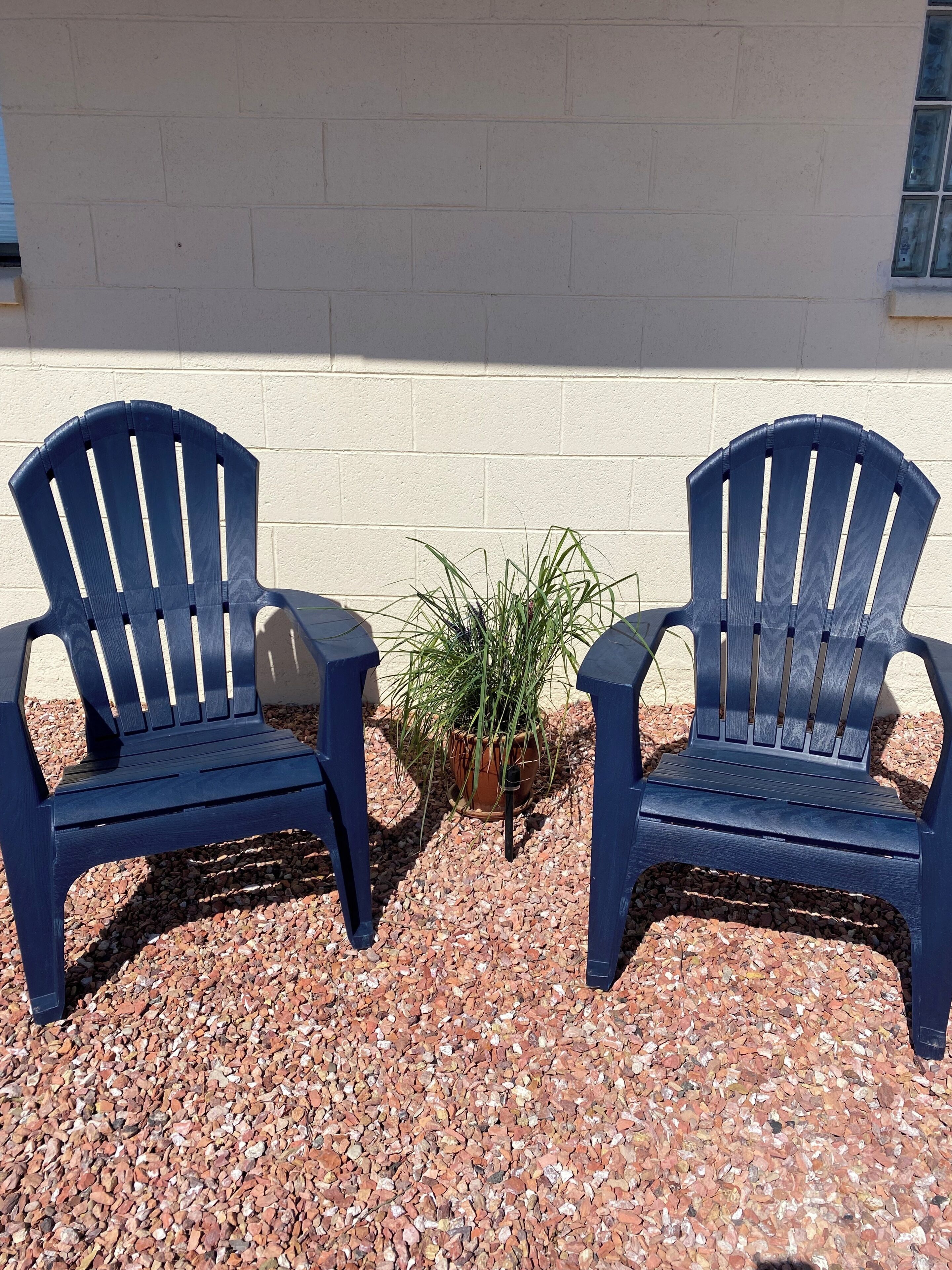 Two blue Adirondack chairs with a potted plant between them, placed on gravel.