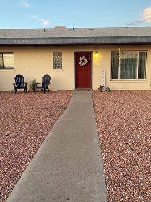 A building with a red door, two Adirondack chairs, a welcome sign, and a pathway leading to the entrance. - Cozy 3-bdrm, 1 bath apt. minutes away from Horseshoe Bend and Antelope Canyon! (Page)