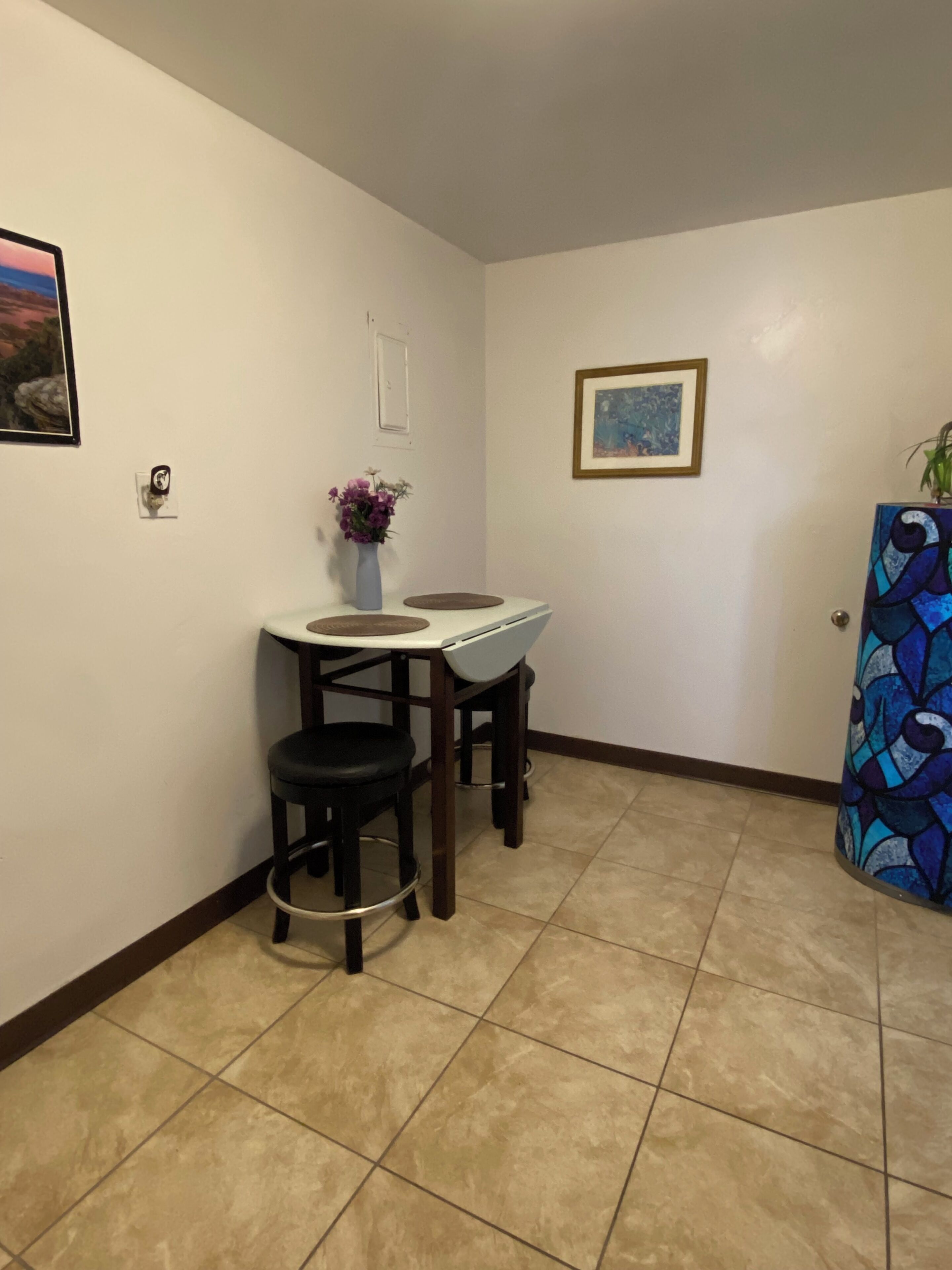A small dining area with a white table and two black stools, a vase with purple flowers, and a framed painting on the wall.