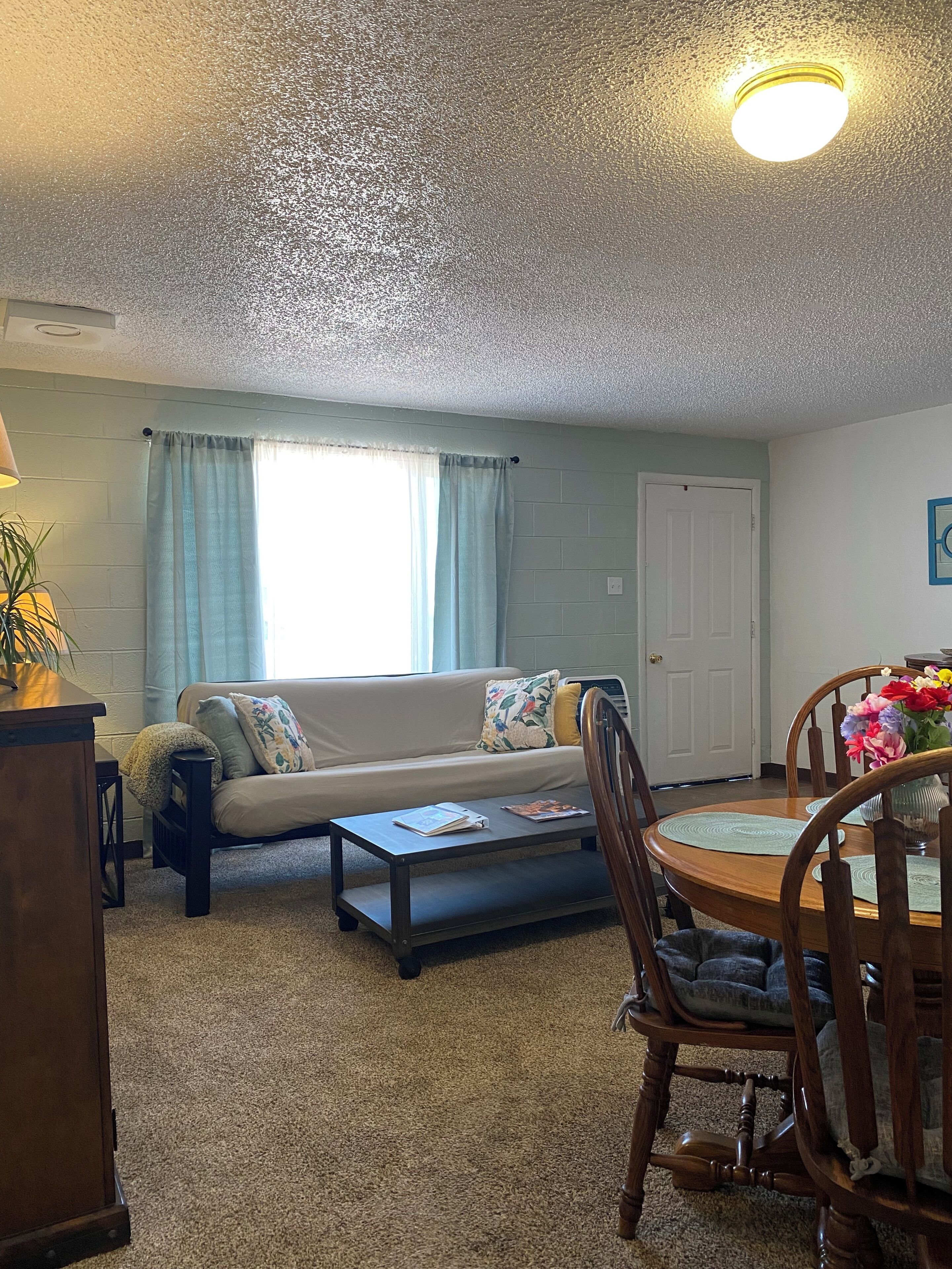A living room with a sofa, coffee table, and dining table with chairs. There is a door and a window with curtains.