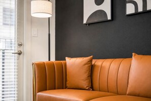 Een leren bank met een kussen, tegen een donkere muur, een vloerlamp en een deur met zonwering.