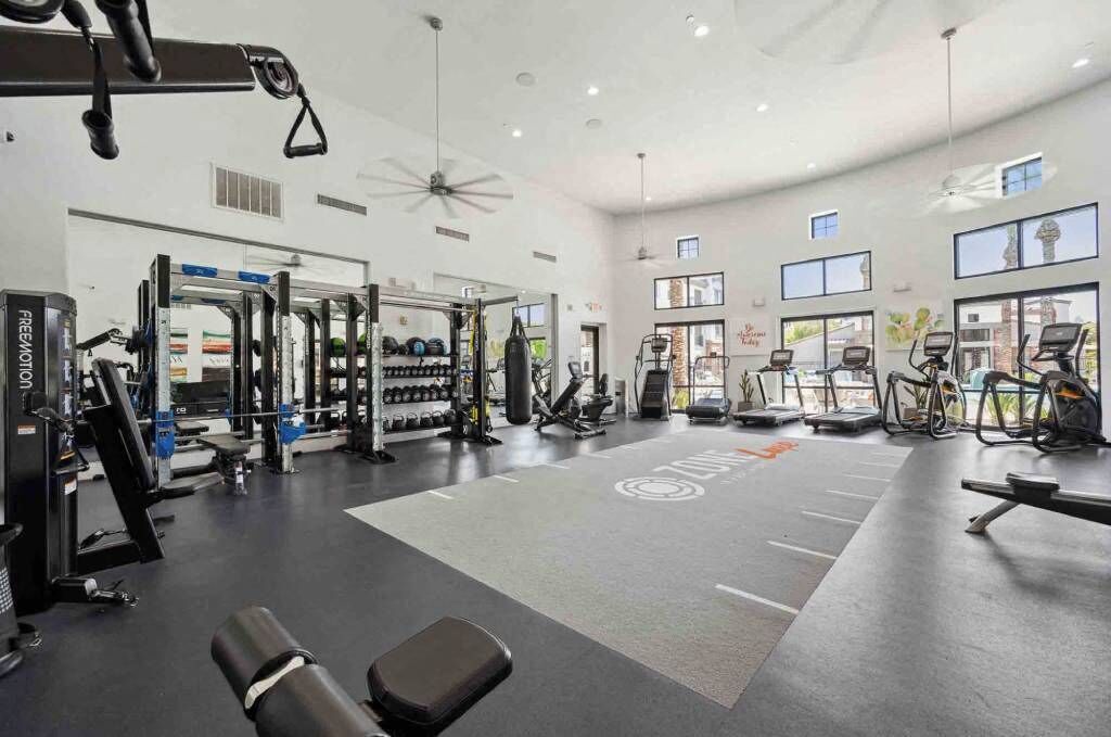 Een ruime fitnessruimte met diverse trainingsapparaten, grote ramen en een plafondventilator.