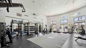 Et rummeligt fitnessrum med forskellig træningsudstyr, store vinduer og en ventilator i loftet.