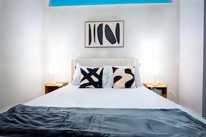 A modern hotel room with a large bed, a white headboard, and a framed abstract artwork above it. There are two bedside tables with lamps. - Boulevard Stays Stylish 1BR Stay Includes Pool, Gym & Free Parking (Dallas)