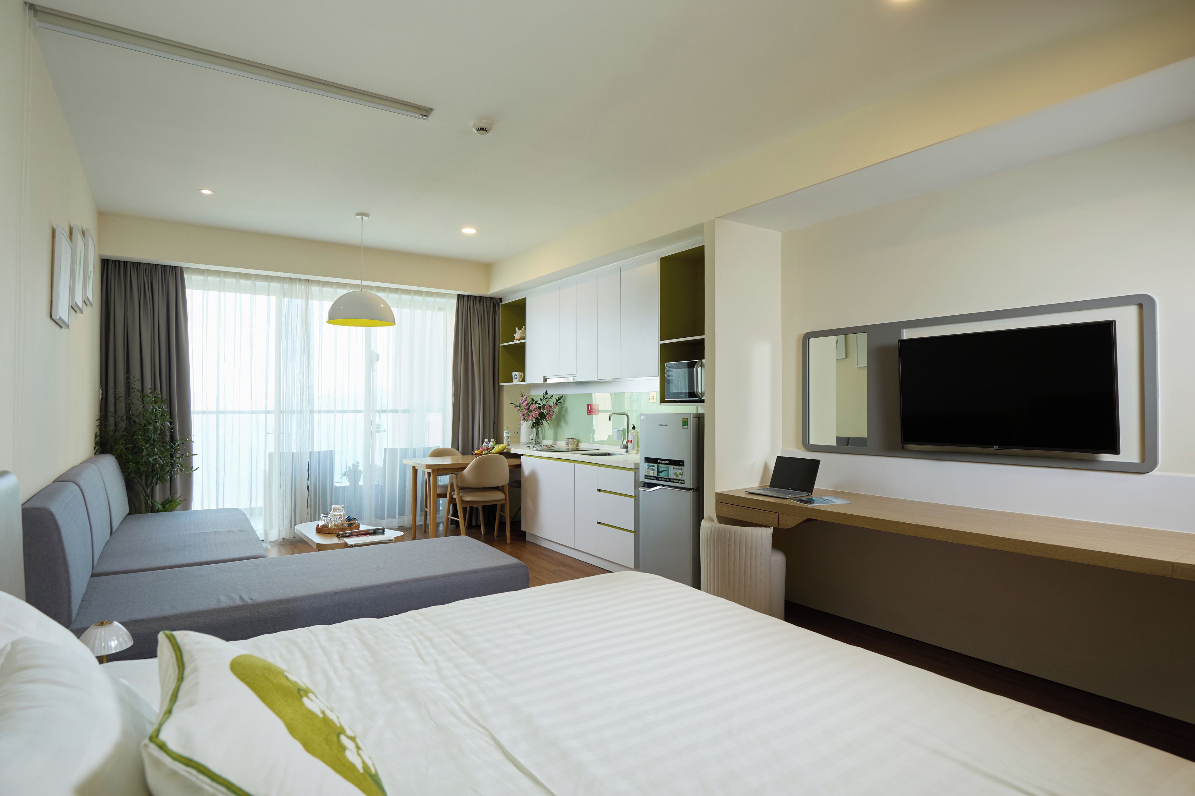 A hotel room with a bed, a desk with a laptop, a kitchenette, and a TV mounted on the wall.