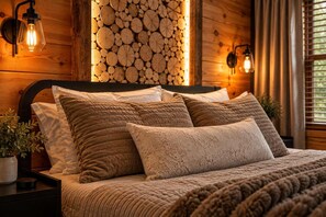 Cama com manta texturizada e várias almofadas, um suporte de madeira fixado na parede e duas luminárias de parede.