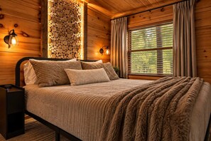 A wooden cabin bedroom with a bed, pillows, a headboard, wall-mounted lights, a window with curtains, and a wall of stacked logs. - Indoor Pool • Hot Tub • Theater • Games • Views (Sevierville)