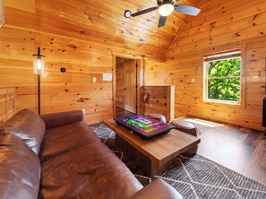 A wooden cabin interior with a sofa, a coffee table, a ceiling fan, and a window with a view of trees. - Indoor Pool • Hot Tub • Theater • Games • Views (Sevierville)
