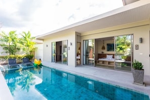A modern poolside area with a clear pool, lounge chairs, and a glass-enclosed room with a bed. - Kaizer Pool Villas (Thalang)