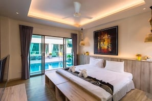 A hotel room with a large bed, a bench, a ceiling fan, a painting, and a view of a pool through a glass door.