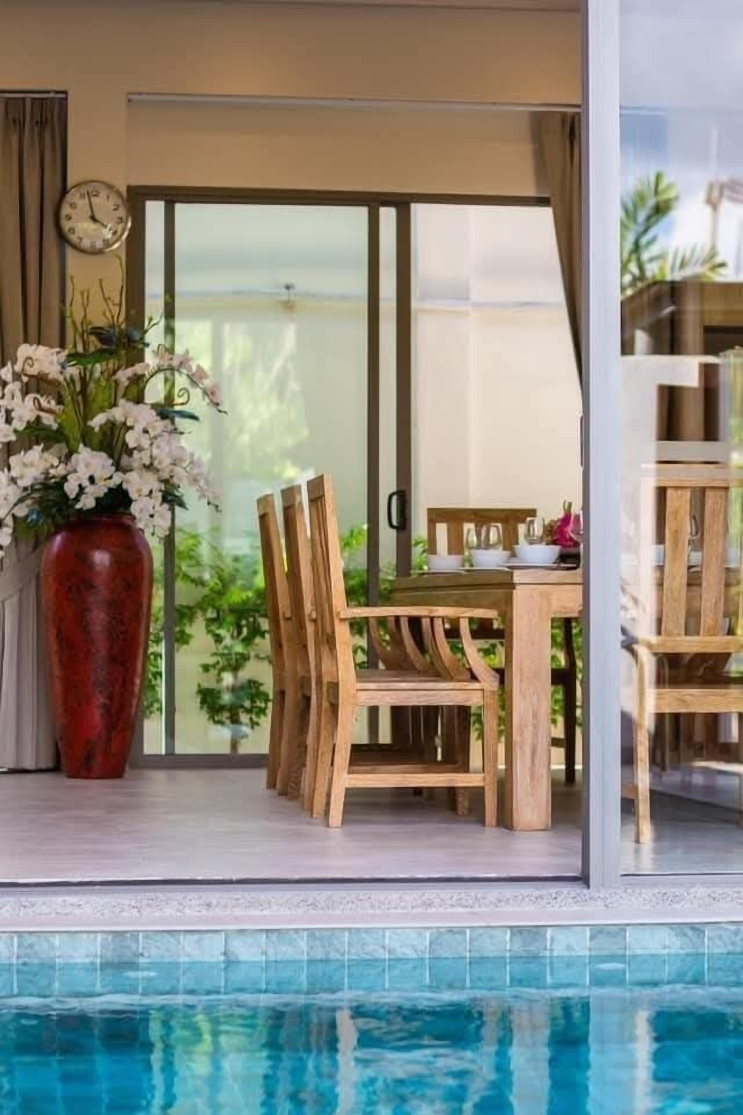 Zona junto a la piscina con una mesa y sillas de madera, un jarrón rojo con flores y vistas al mar a través de un ventanal.