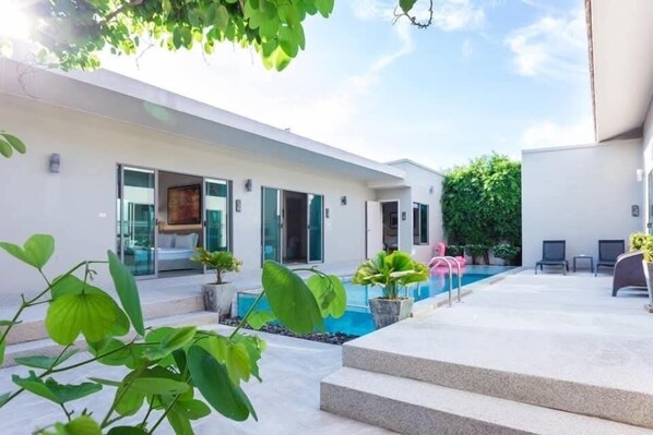 A modern outdoor pool area with steps leading to a glass door entrance, surrounded by potted plants and a clear sky.