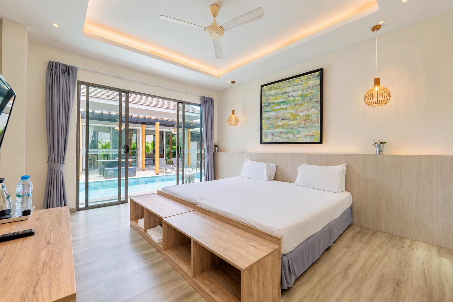 Une chambre d’hôtel moderne avec un grand lit, un banc en bois, un bureau, un ventilateur de plafond et une vue sur la piscine, visible à travers des portes vitrées.