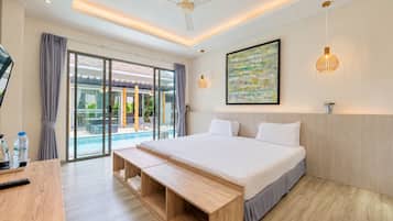 Une chambre d’hôtel moderne avec un grand lit, un banc en bois, un bureau, un ventilateur de plafond et une vue sur la piscine, visible à travers des portes vitrées.