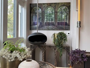 A room with a large window, a painting of an arched window, a black object on a stand, and various potted plants. - Spacious Modern Apartment in the heart of Fitzroy (Melbourne)