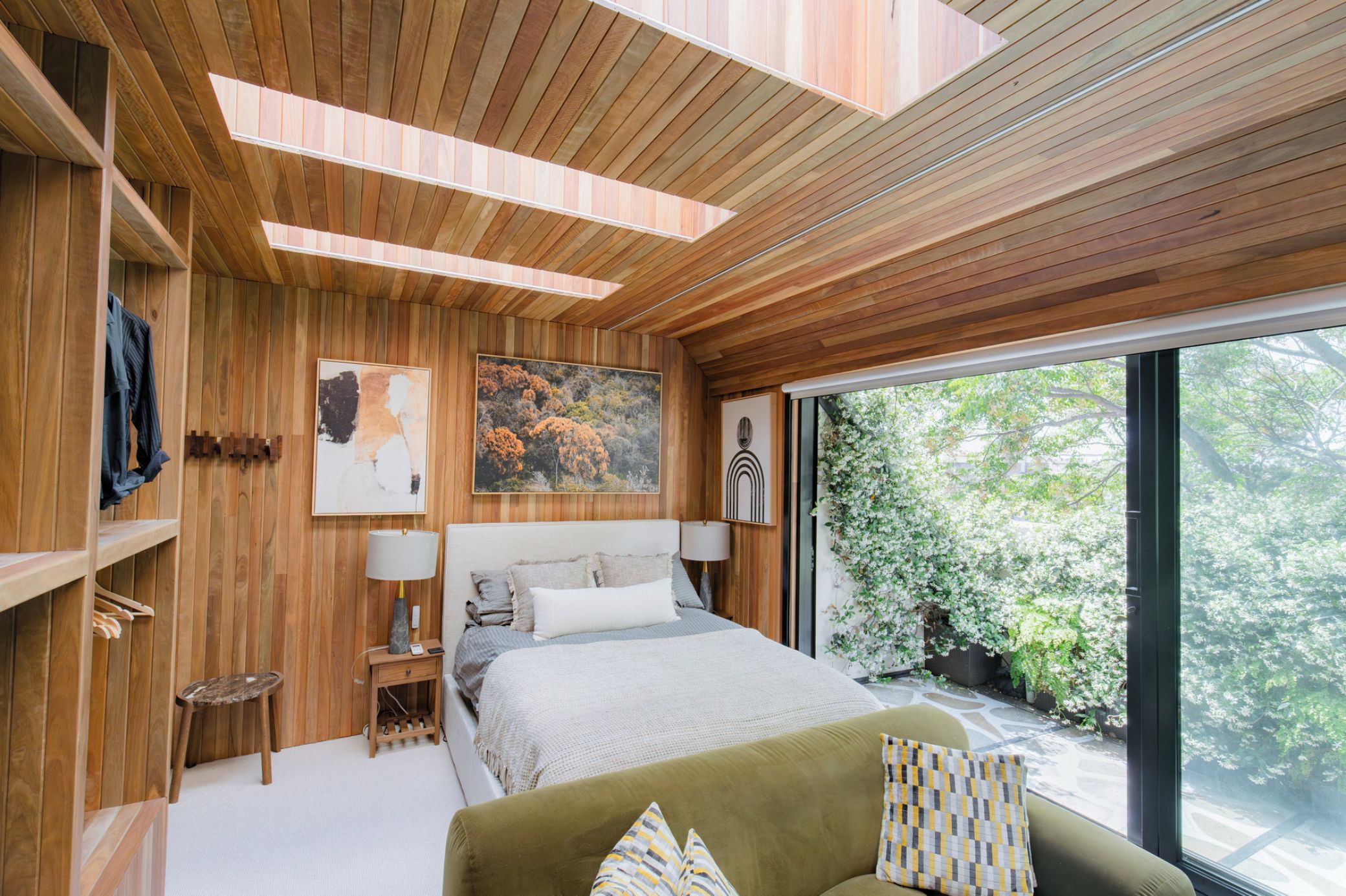 A modern bedroom with a large bed, a green sofa, wooden panel walls, and a view of greenery outside.