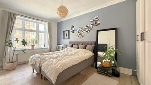 A bedroom with a large bed, a window with curtains, a wall-mounted mirror, and a potted plant.