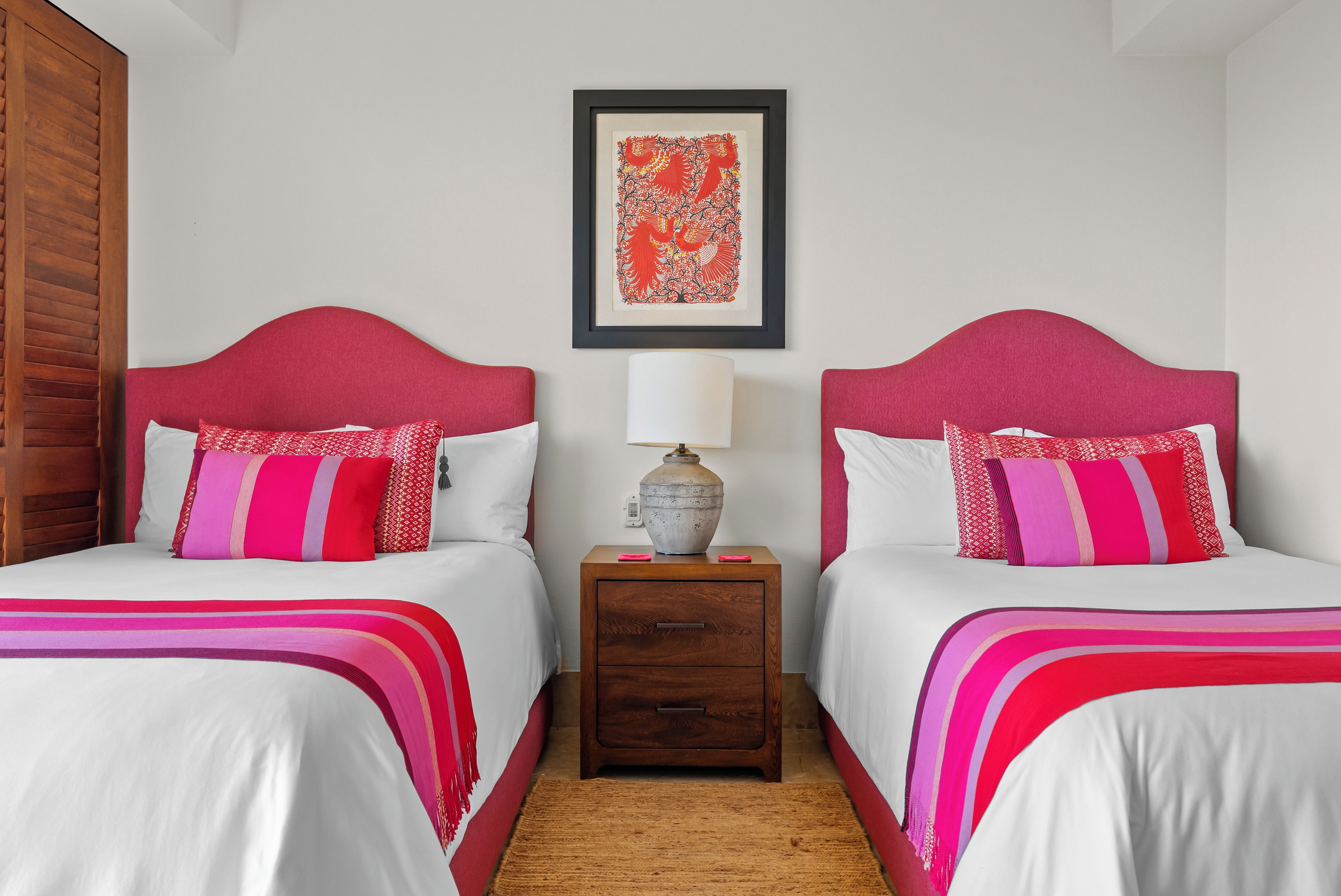 A hotel room with two beds, each with a red headboard, white bedding, and pink and white patterned pillows. There is a nightstand with a lamp and a decorative vase between the beds.