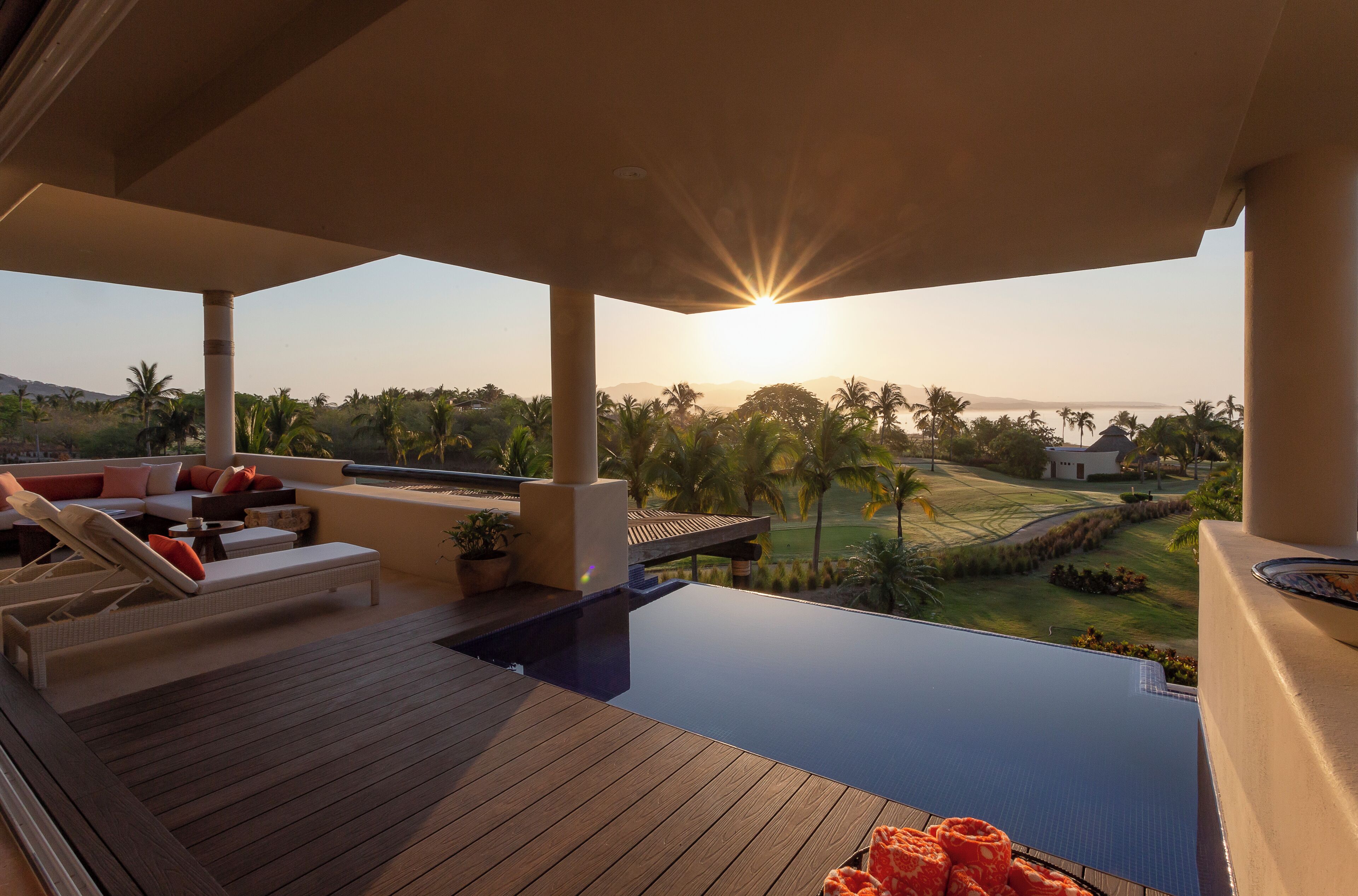 Une piscine à débordement baignée de soleil, avec une terrasse en bois, un espace détente doté de coussins et une vue sur un terrain de golf et des palmiers.
