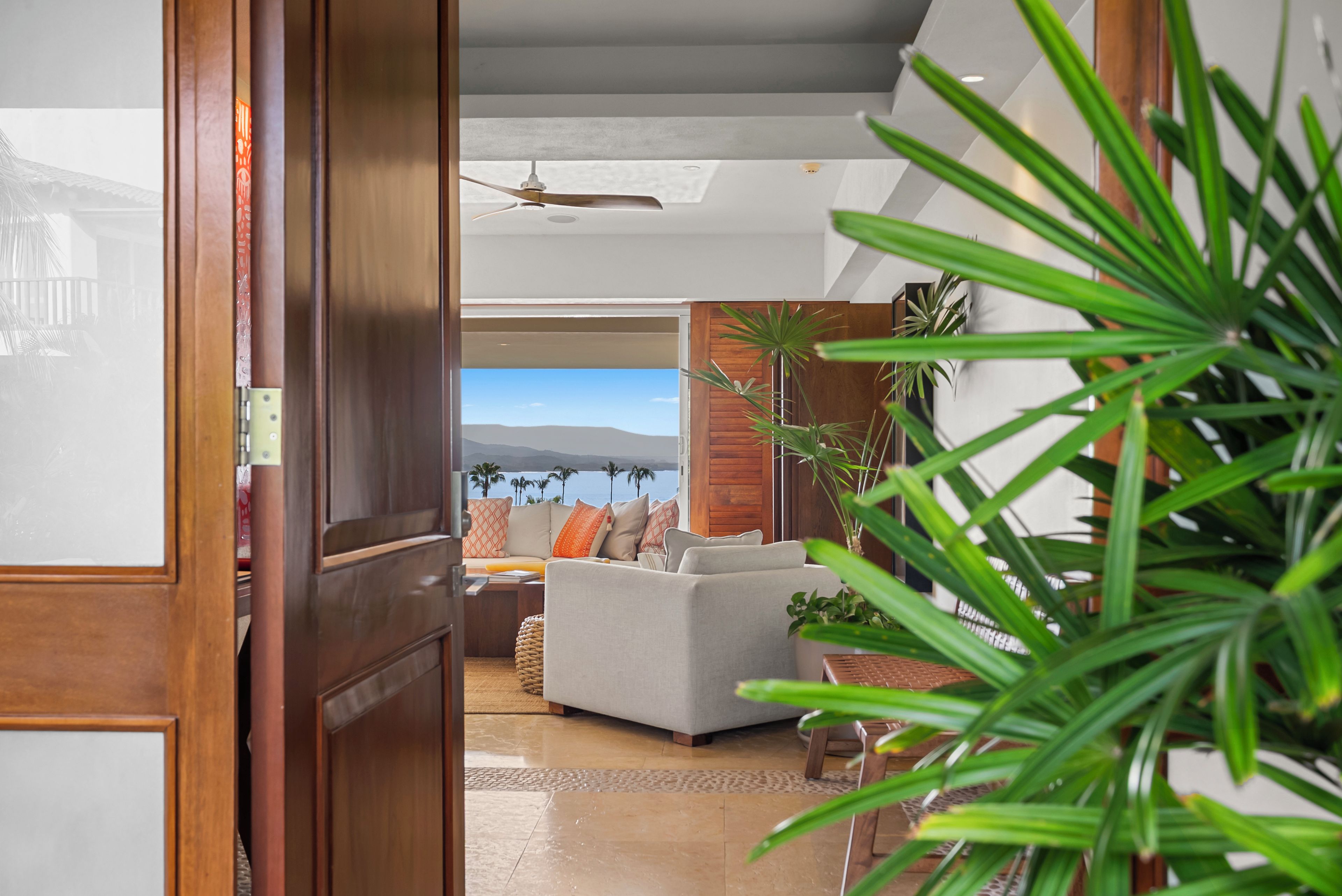 Ein Blick ins Innere eines Zimmers mit Holztür, Deckenventilator, einer Couch mit Kissen, einem Couchtisch und Blick ins Freie.