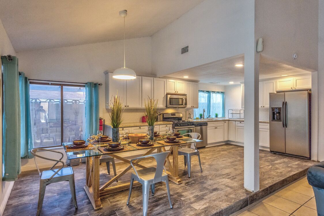 A modern kitchen with white cabinets, a stainless steel refrigerator, and a dining area with a glass table and metal chairs.
