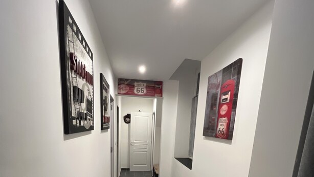 A hallway with dark tiled flooring, white walls, and framed pictures on the walls. There is a door at the end of the hallway. - -In Valjc- (Liverdun)