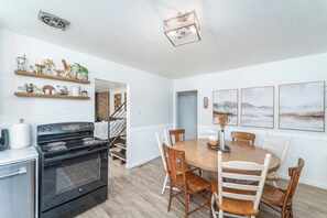 Een keuken met een fornuis, oven en planken met diverse spullen. Er is een eettafel met stoelen en een trap naar een andere kamer.