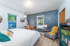 Een slaapkamer met een bed, een bureau, een stoel, een boekenplank en een raam met uitzicht op bomen.