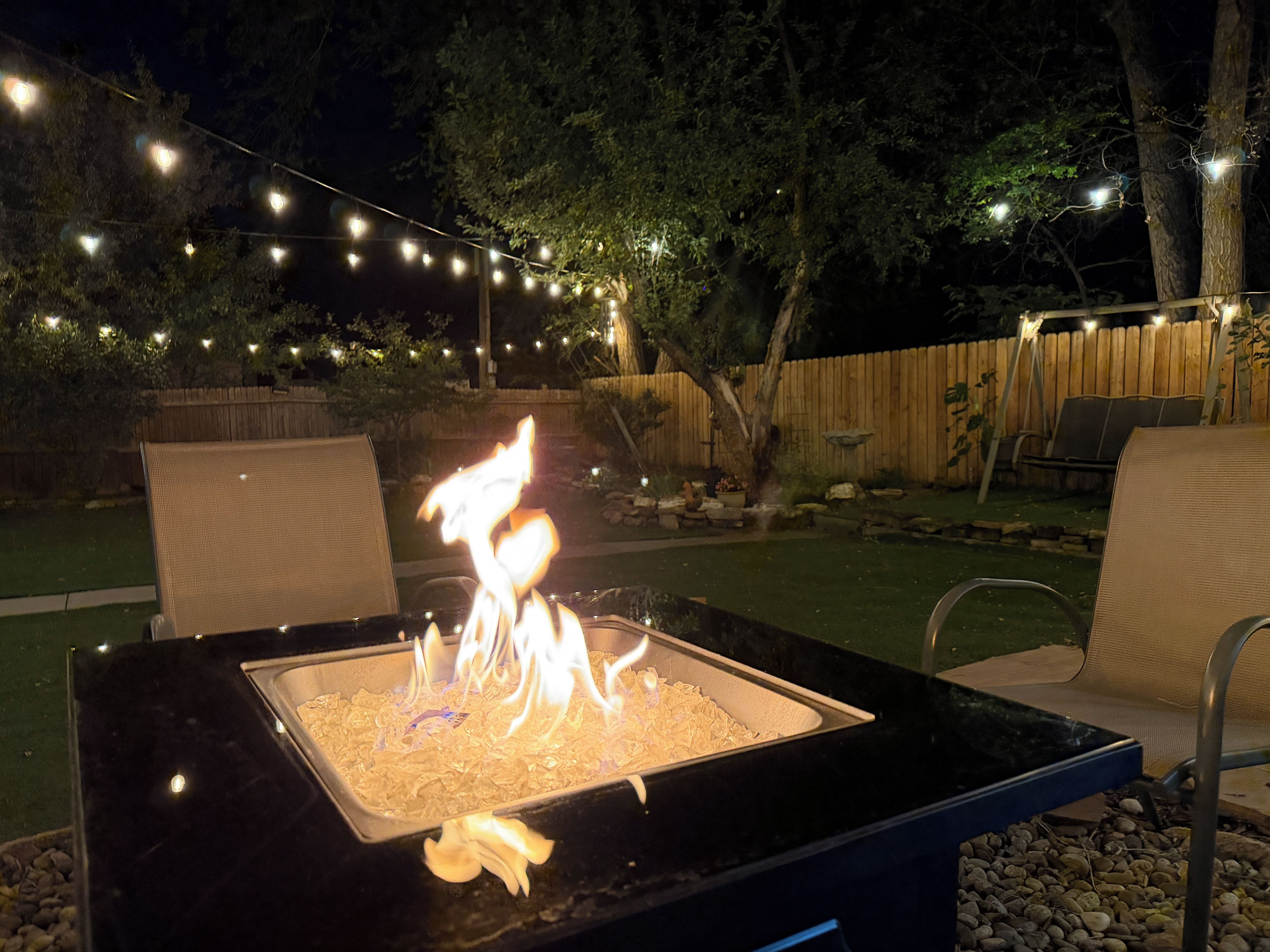 An outdoor fire pit with flames, surrounded by chairs and a string of lights.