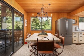 Un intérieur en bois comprenant une table à manger, des chaises, un lustre, un réfrigérateur et une vue dégagée sur l’extérieur grâce à de grandes fenêtres.
