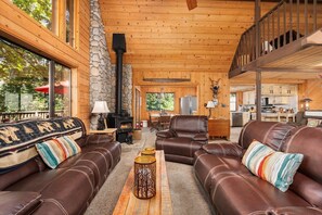 A log cabin interior with leather sofas, a stone fireplace, and a kitchen in the background. - Wyldwood Cabin | Pet Friendly | Shaver Lake (Shaver Lake)
