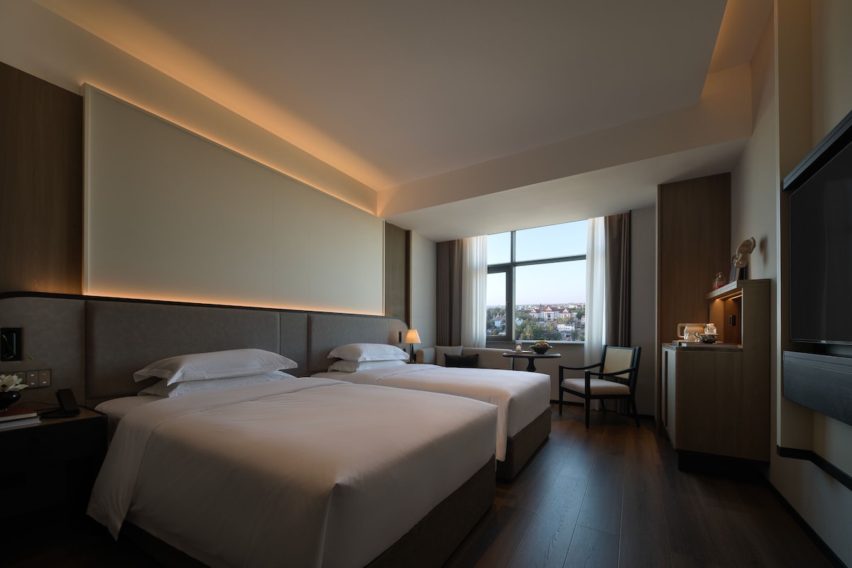 Hotelkamer met twee bedden, een bureau, een stoel, een televisie en een raam met uitzicht over de stad.