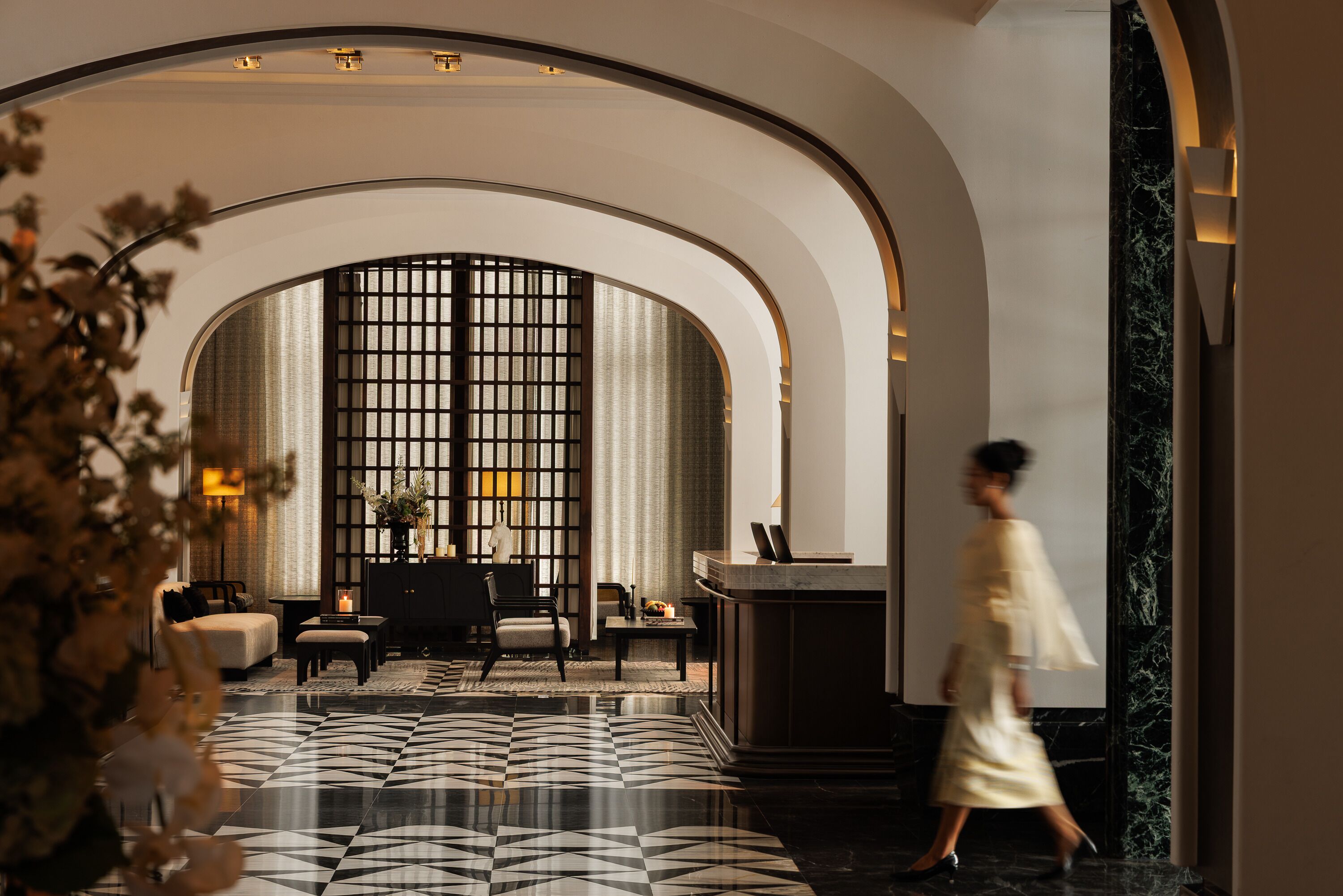 A hotel lobby with a geometric patterned floor, arched ceiling, and a seating area with a view through a grid-like window.