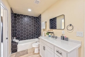 Ein Badezimmer mit Wand aus sechseckigen Fliesen, einer Badewanne, einer Toilette, einem Waschbecken mit Spiegel und einem Handtuchhalter.
