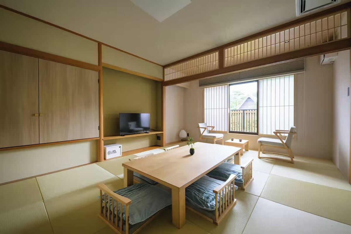 Quarto em estilo tradicional japonês, com tatames, uma mesa baixa e uma televisão em uma estante.