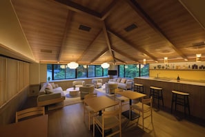 A modern interior with a high ceiling, wooden beams, a seating area with sofas and tables, a kitchen with bar stools, and large windows offering a view of greenery.