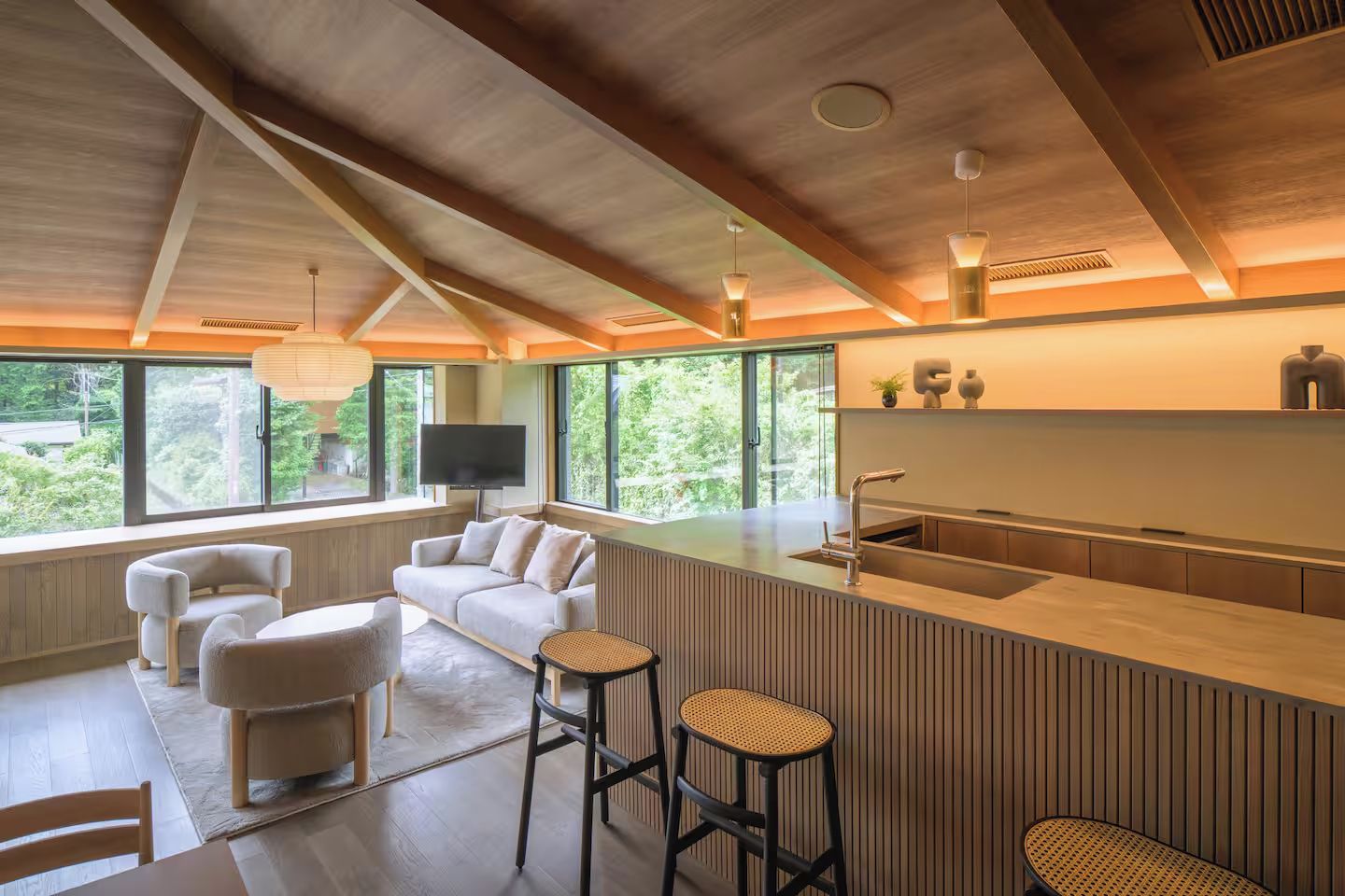A modern kitchen with a bar area, a seating area with a sofa and chairs, and large windows offering a view of greenery.
