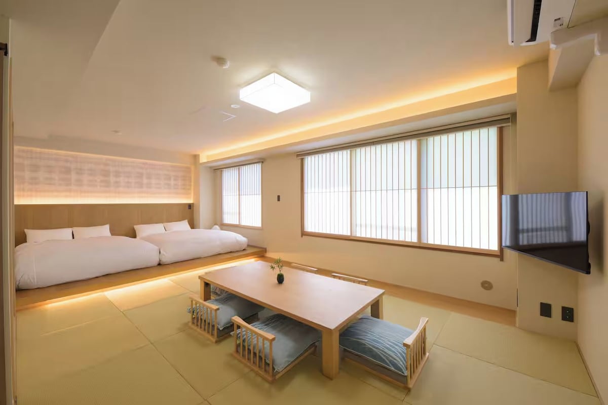 Quarto de hotel moderno com sofá-cama, mesa de jantar e televisão fixada na parede.