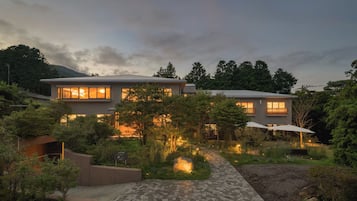 A modern two-story house with a stone pathway leading to the entrance, surrounded by lush greenery and a pond.